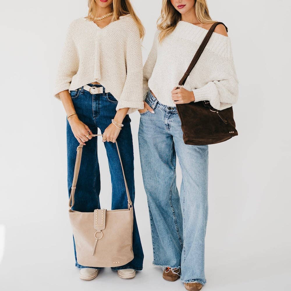Two women modeling off-the-shoulder knit sweaters and jeans, each carrying a large slouchy crossbody bag. One bag is light beige and the other is dark brown, shown against a clean white studio background.