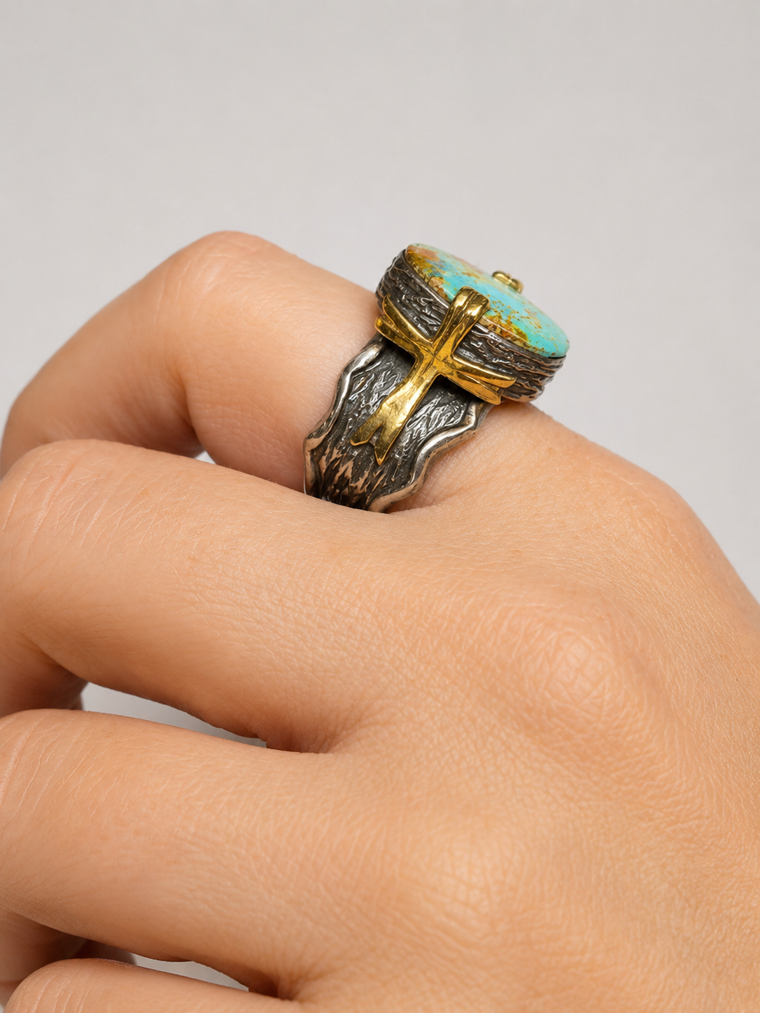 A close-up of a hand wearing a rustic silver ring with a turquoise and gold cross-shaped stone.