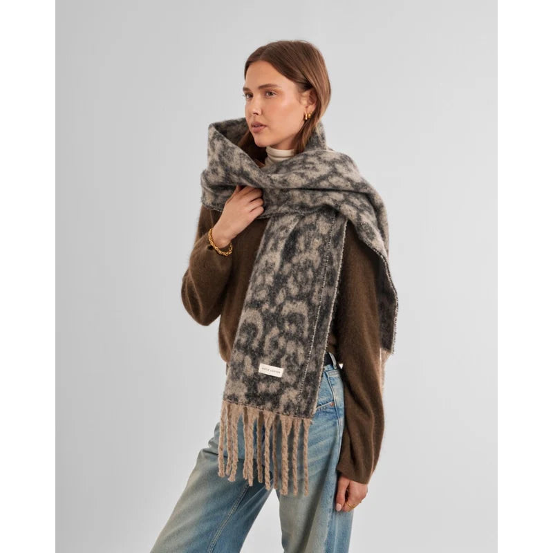 A woman modeling a thick, soft knit scarf with a mottled gray and beige animal-like pattern and fringed ends, styled over a brown sweater and blue jeans against a light gray studio background.