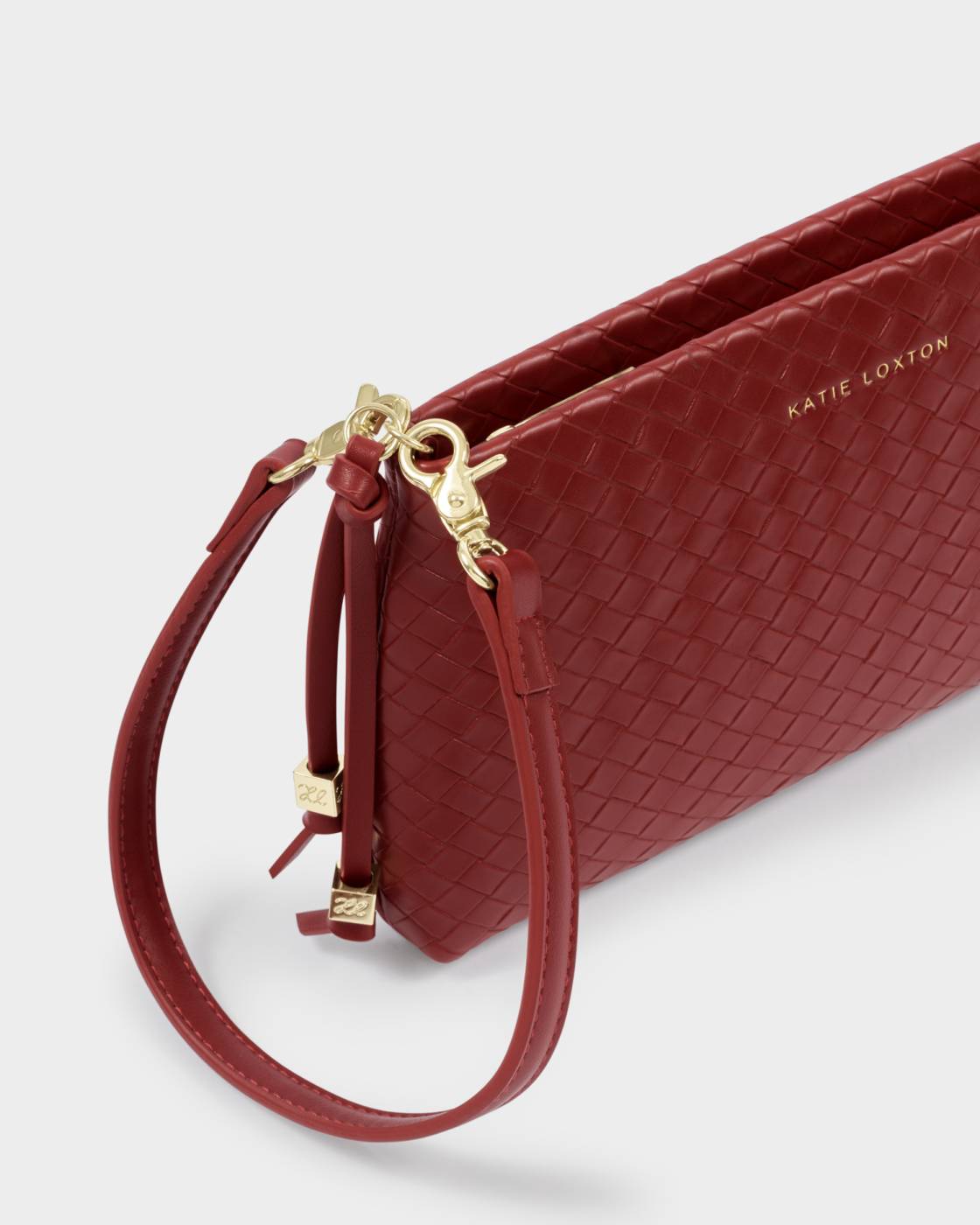 A close-up of a red woven leather clutch with a wristlet strap and gold-tone hardware, featuring the brand name 'KATIE LOXTON' on the side, set against a light gray background.