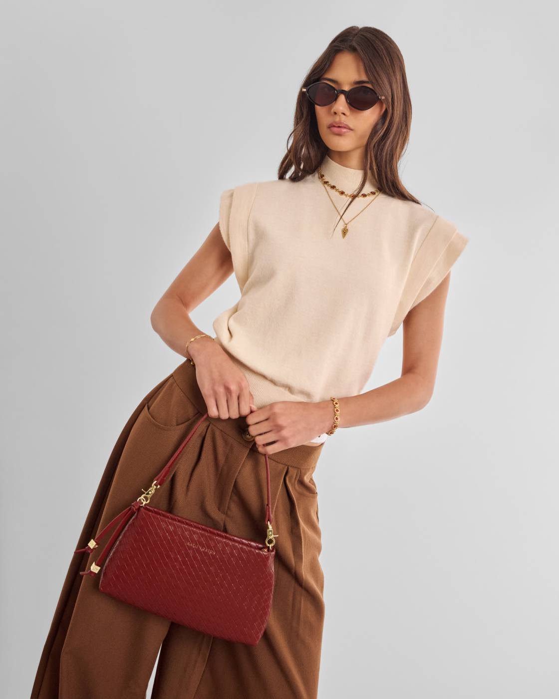 A model wearing a cream-colored sleeveless knit top, brown wide-leg trousers, gold jewelry, and sunglasses, holding a textured dark red handbag with gold hardware against a light gray studio background.