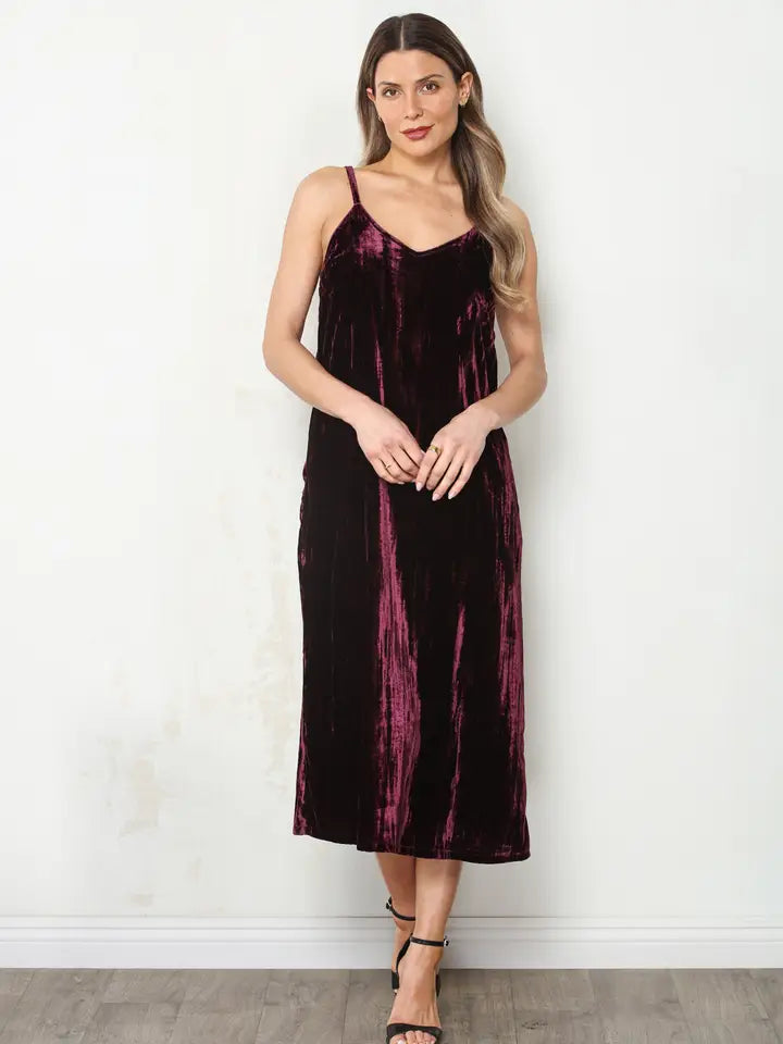 A woman modeling a sleeveless midi slip dress in a deep plum velvet fabric, standing against a plain off-white wall.