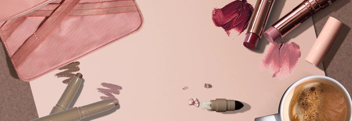 Merle Norman lipsticks, cream eyeshadow sticks, a blush pink makeup bag and a latte on a warm pink background