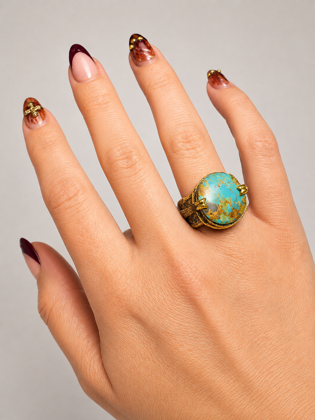 A close-up of a hand wearing a gold ring with a large oval turquoise stone, set against a plain light gray background.