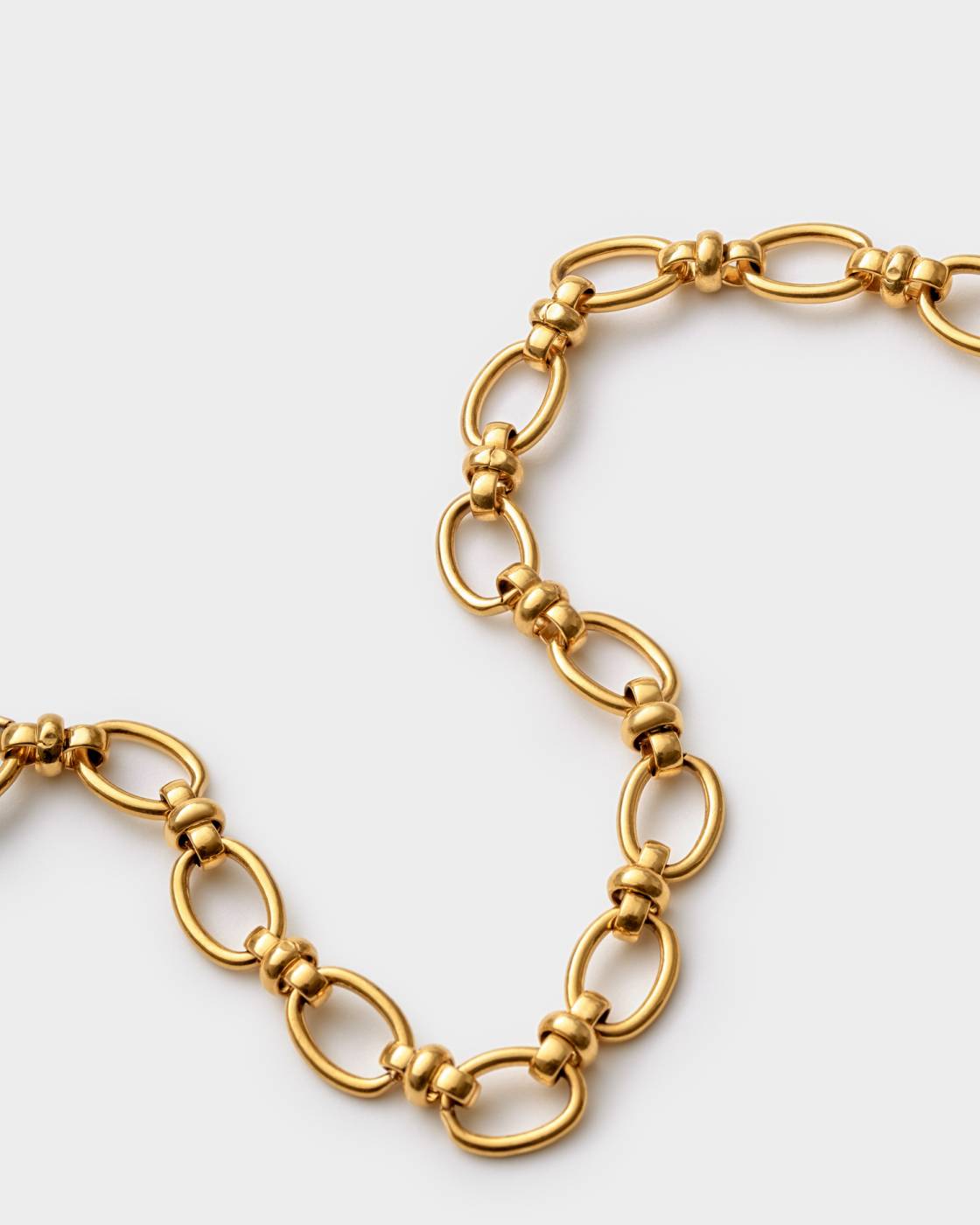 A close-up of a gold-toned chain necklace with elongated oval links and smaller beaded connector links, photographed on a light gray background.