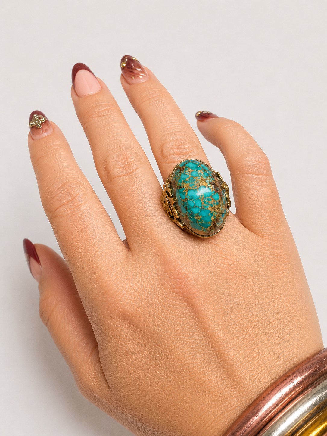 A close-up of a hand wearing a large oval turquoise ring with gold detailing, paired with stacked copper and gold bangles, against a light gray background.