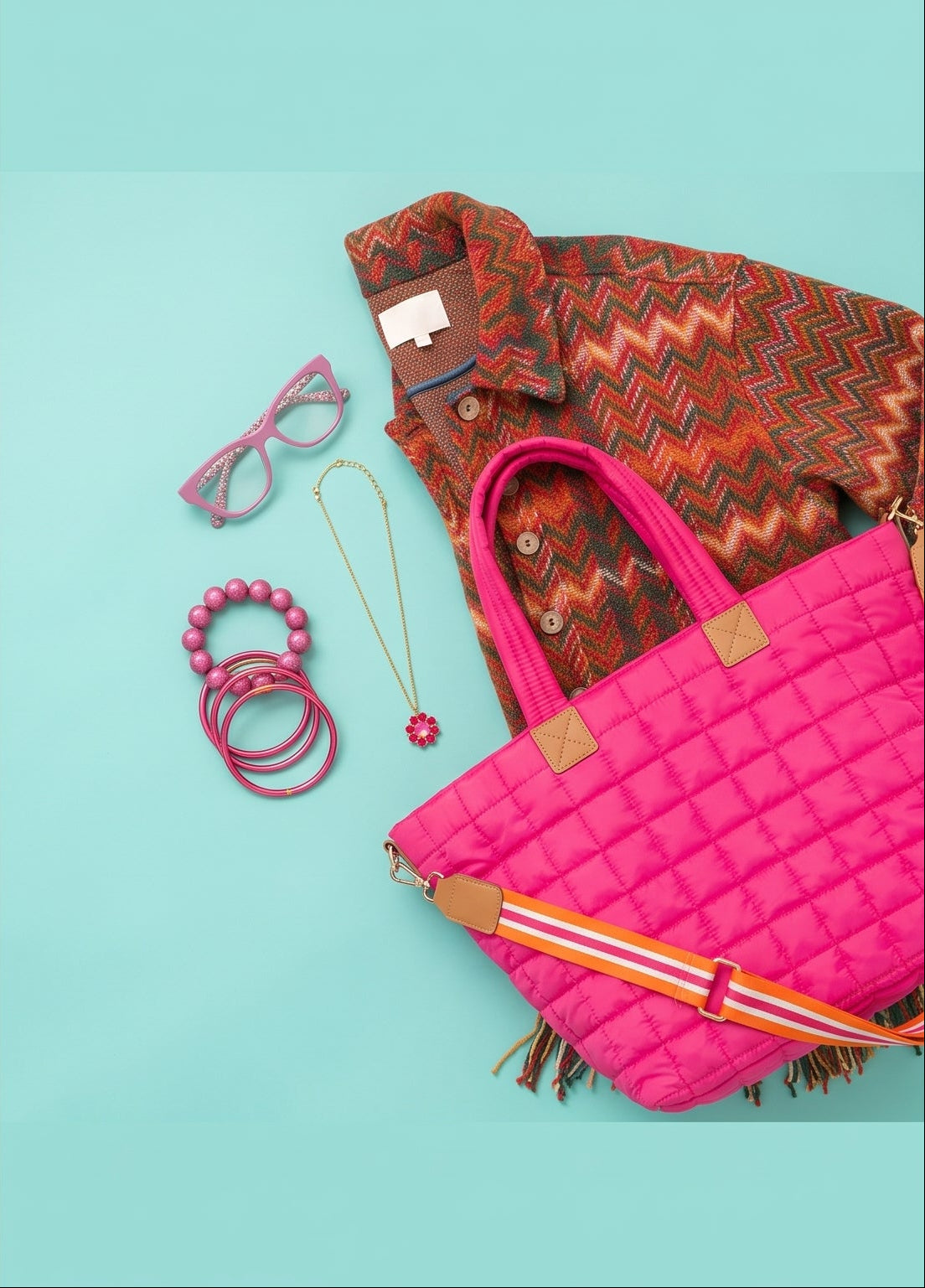 Flat-lay of Pink Out pieces on a turquoise background: a hot pink quilted tote with a striped strap, stacked pink bangles and a beaded bracelet, pink reading glasses, a gold chain necklace with a pink floral pendant, and a chevron knit jacket