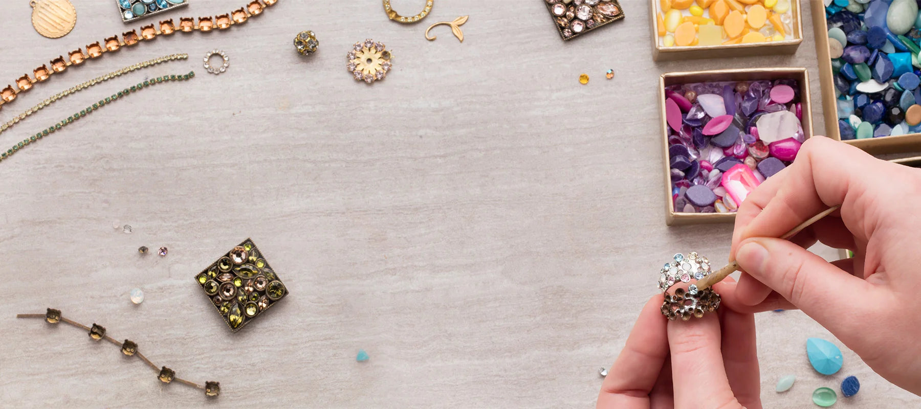 A close-up of a person's hands assembling a beaded jewelry component on a light gray wooden surface, surrounded by various colorful beads, sequins, and jewelry-making supplies.