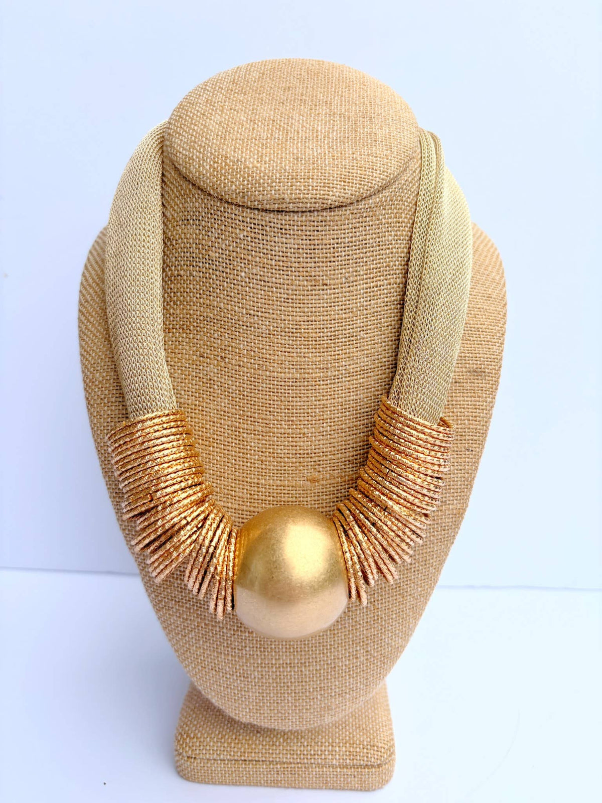 A chunky statement necklace with a matte gold central sphere and textured gold fringe, displayed on a beige fabric jewelry bust against a light blue background.